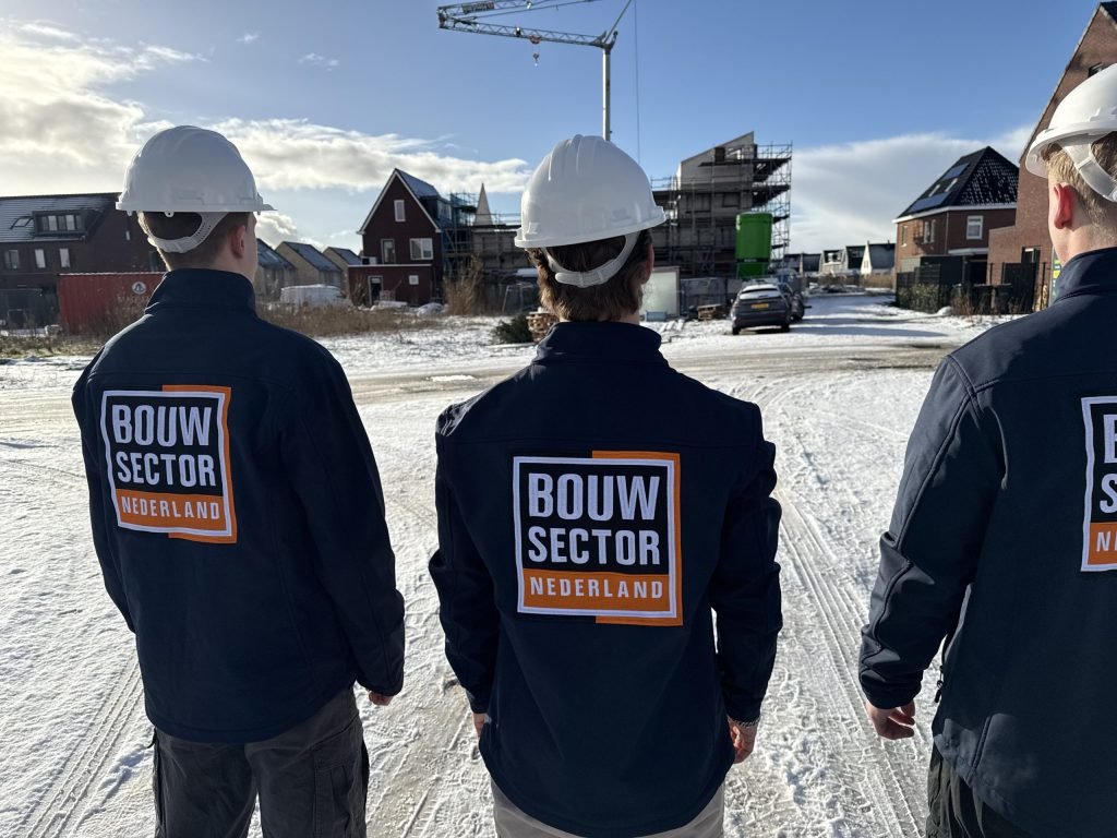 Drie medewerkers van Bouwsector Nederland in donkerblauwe jas en witte helm op een bouwterrein met nieuwe huizen